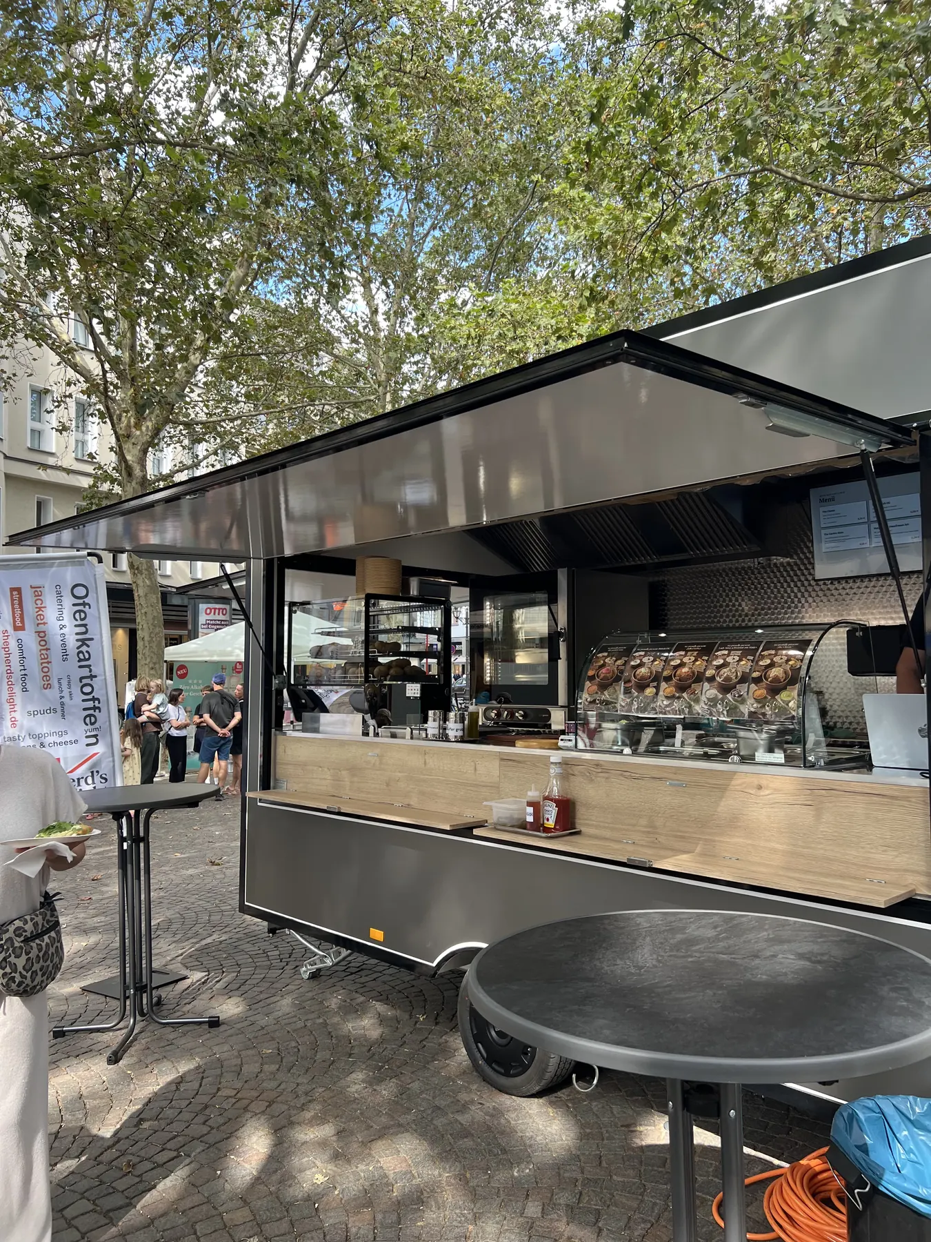 Foodtrailer mit geöffneter Ausgabe, tagsüber auf einem Platz mit Bäumen