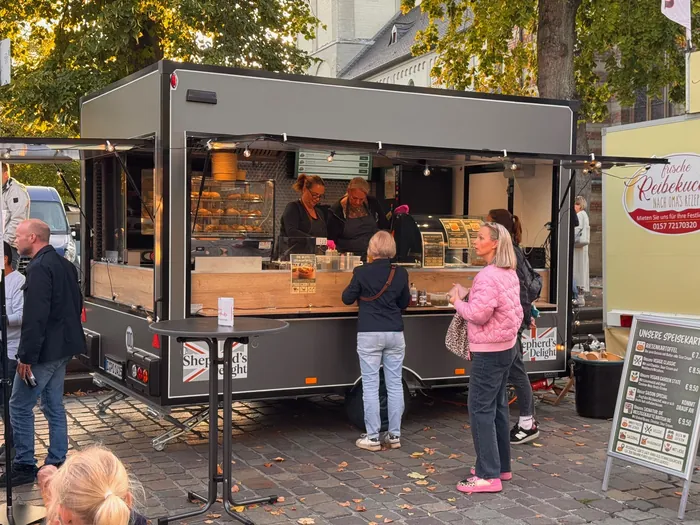 Shepherd's Delight Foodtrailer bei Büttgen kulinarisch: geöffnete Ausgabe mit Lichterkette in der Abendsonne auf dem Kirchplatz, Gäste an der Theke